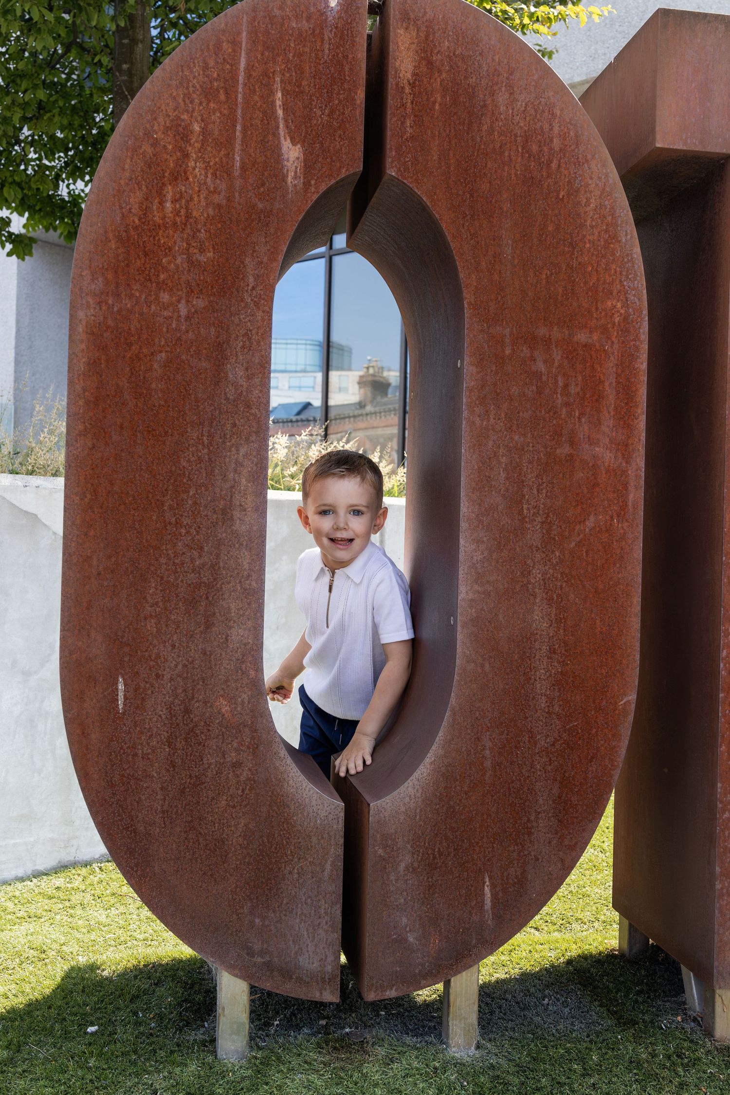 Young boy smiling and posing inside a large rusty metal number zero sculpture outdoors on green grass.