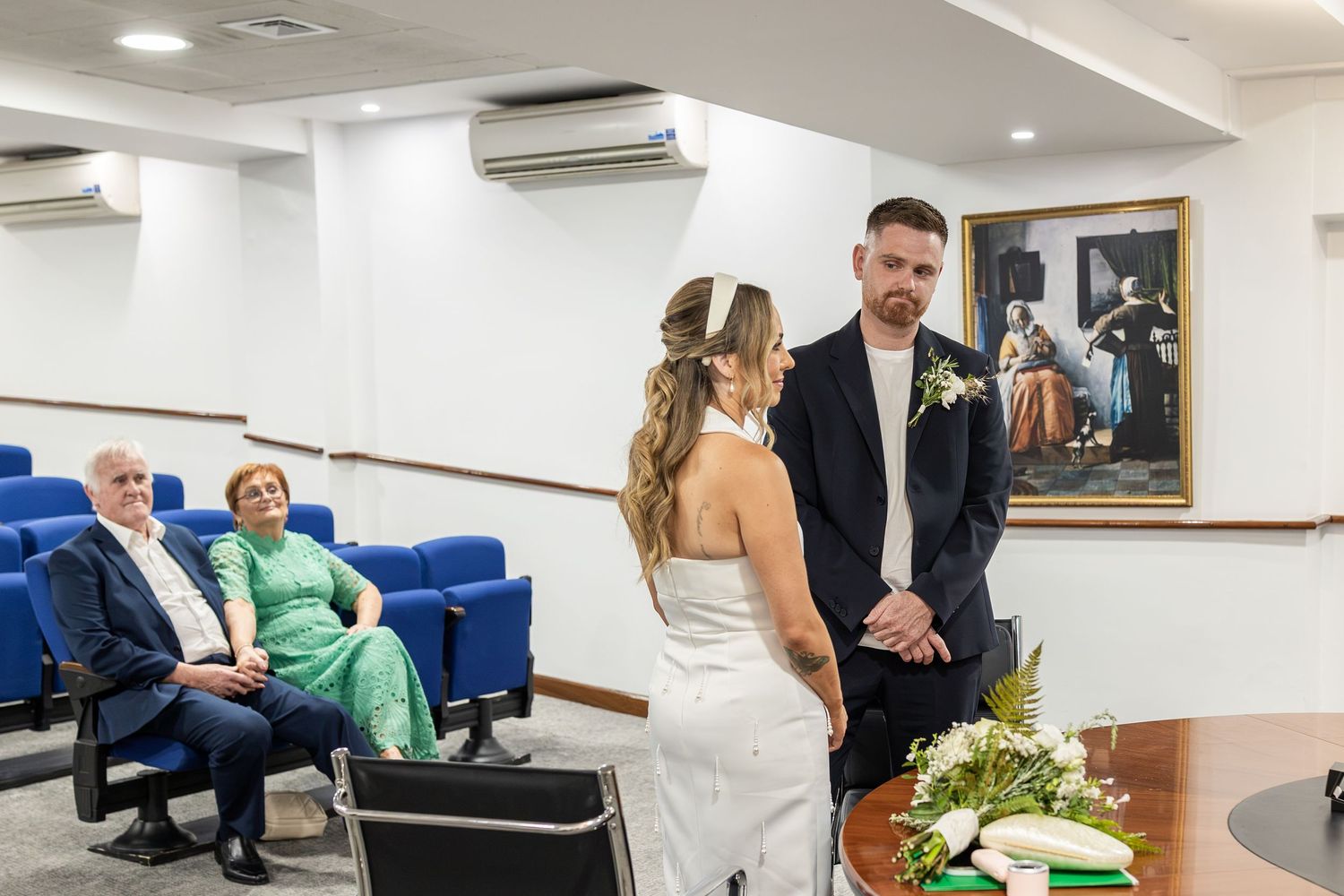 Couple exchanges vows at intimate civil wedding ceremony with guests seated in blue chairs in a formal registry office.