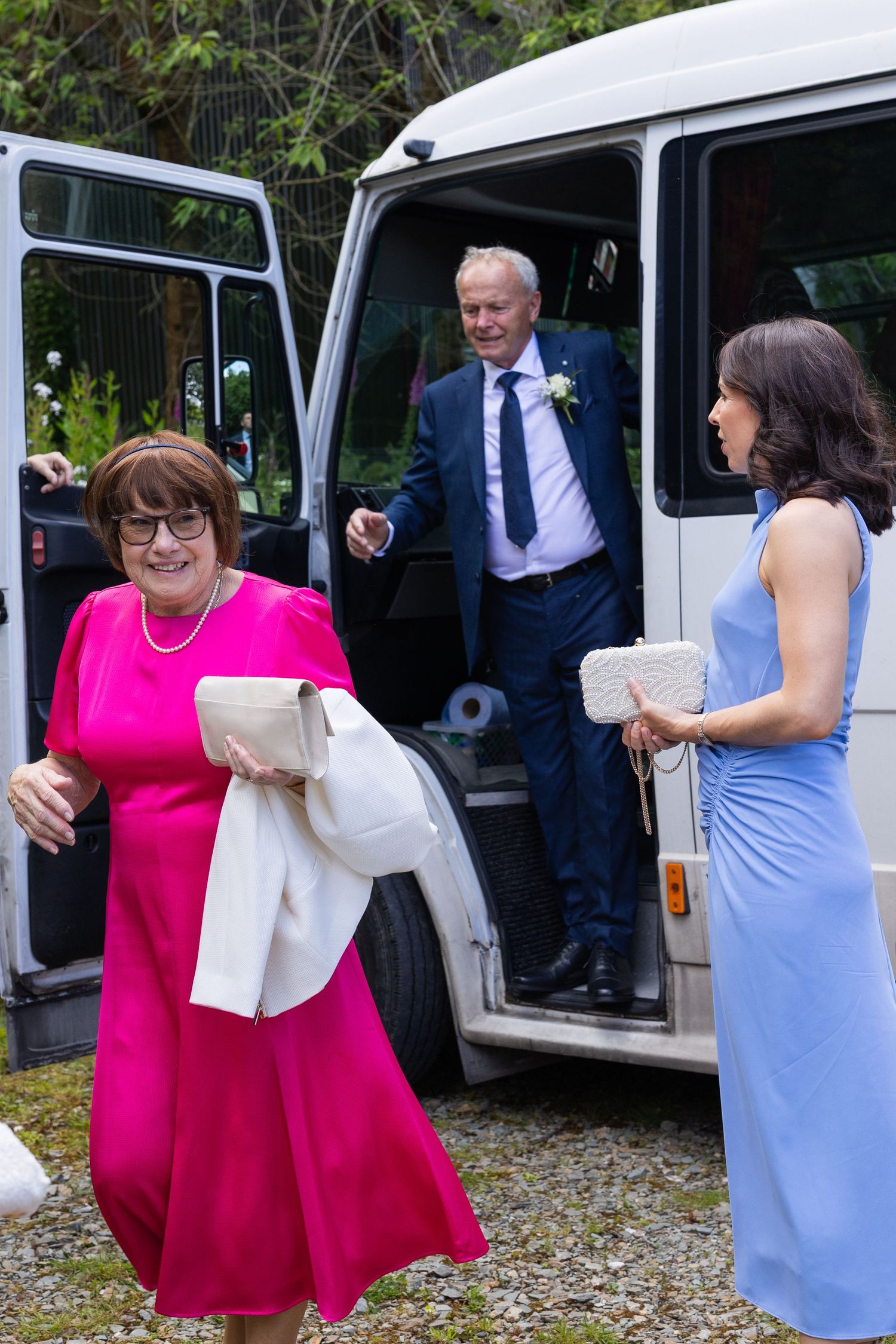 Wedding guests in formal attire exit a white minibus, woman in pink dress and man with boutonniere smiling.