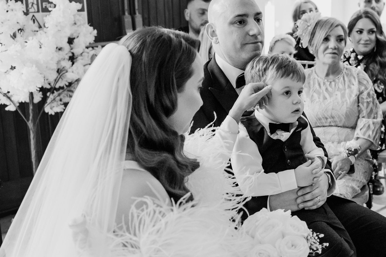 Black and white wedding photo of bride and groom seated with young boy in bow tie during ceremony.