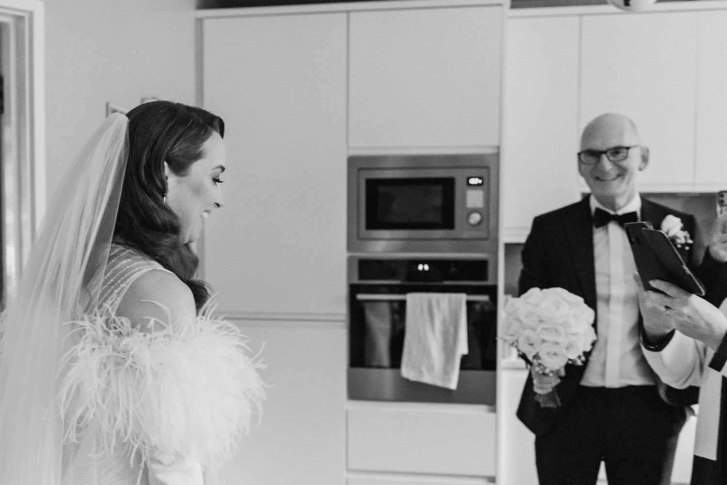 Bride in feathered wedding dress shares first look moment with father in tuxedo holding bouquet.