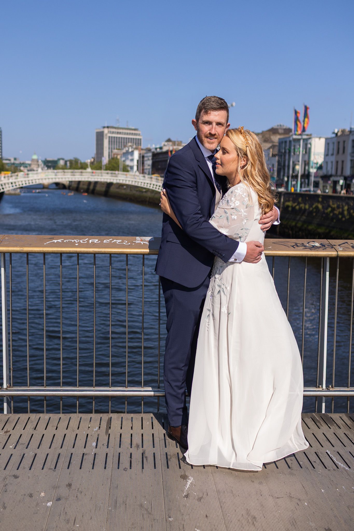 A wedding couple embraces on a waterfront boardwalk with ocean views in the background.
