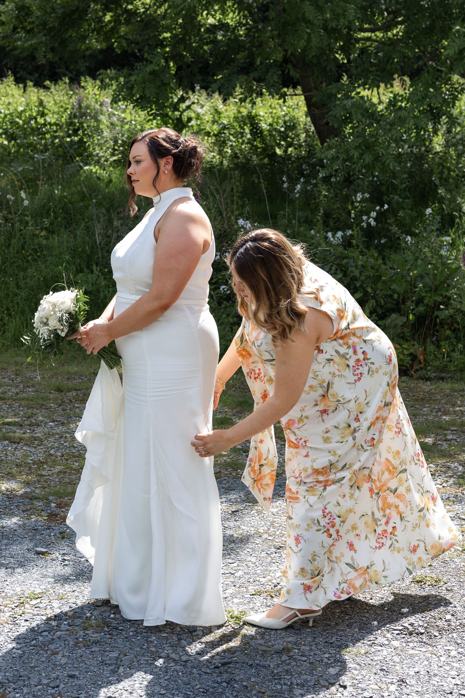 Bridesmaid in floral dress adjusting bride's white halter wedding gown train outdoors on gravel path.