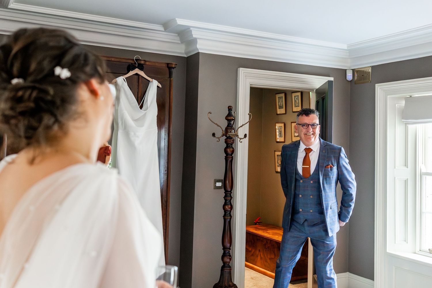 Groom in blue suit smiles at bride during wedding morning preparations, with bridal gown hanging in background.