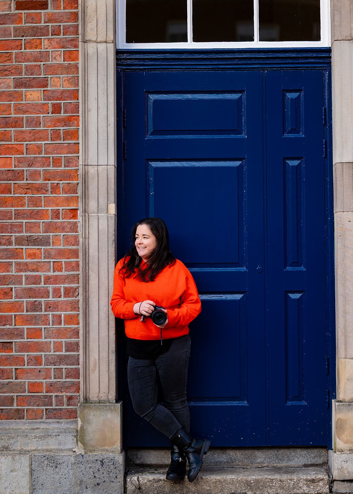 Person in bright orange jacket posing against blue doors and brick wall architecture.