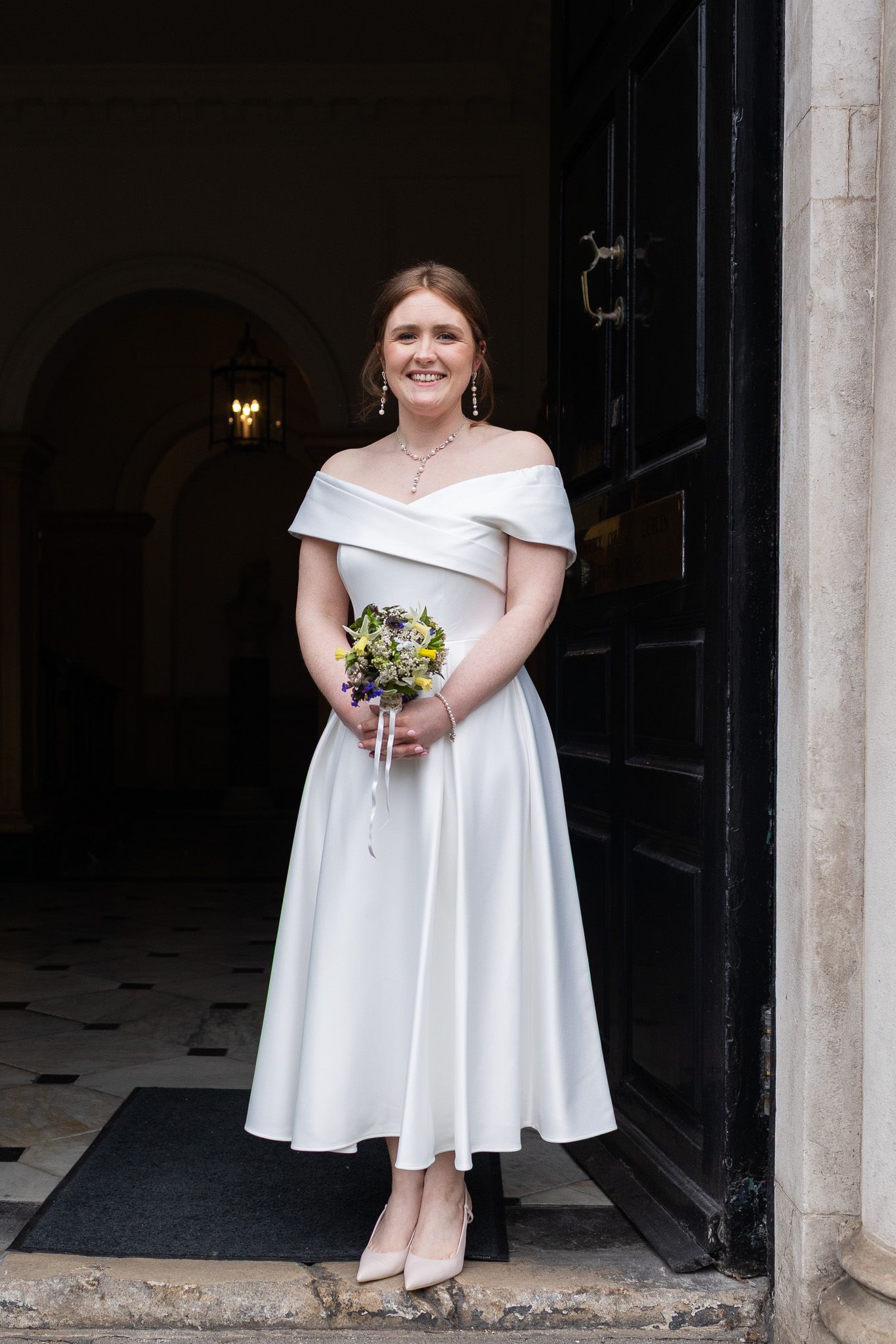 Bride in white dress holding bouquet stands in elegant MoLI  doorway entrance during Dublin City intimate wedding.