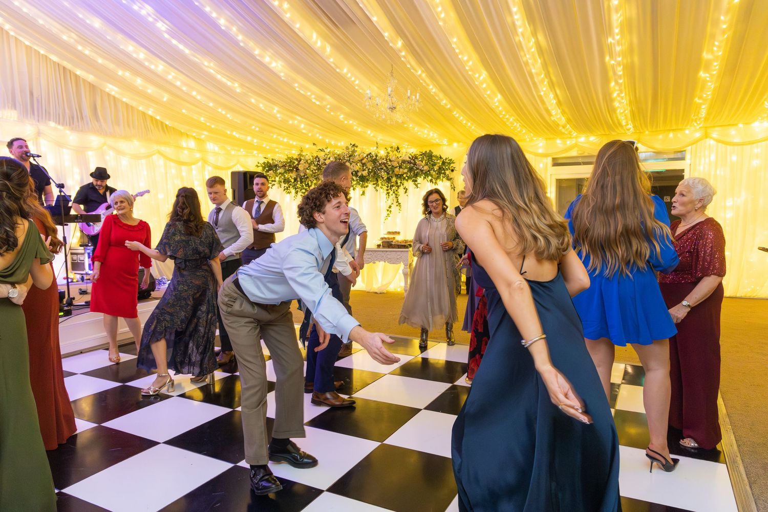 Guests dancing on checkered floor at wedding reception under warm golden fairy light canopy.