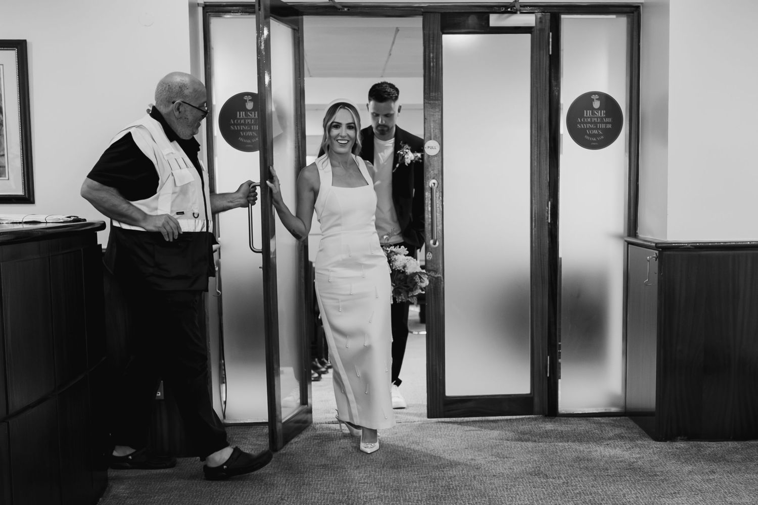 Black and white photo of a bride in white gown entering a building through glass doors with groom and usher.