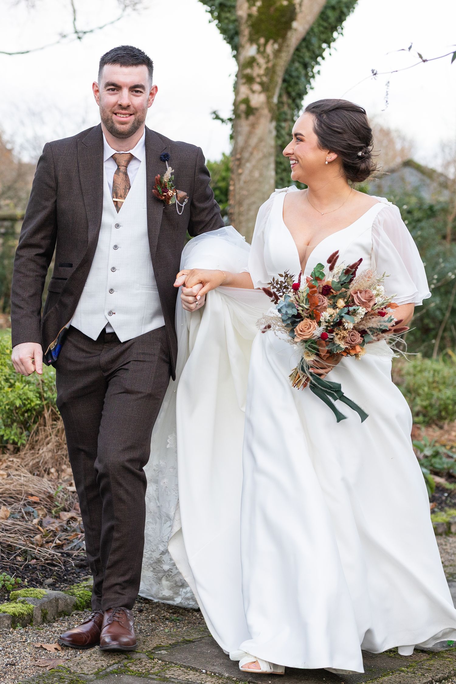 A couple poses in formal wedding attire against a natural outdoor backdrop with autumn foliage.