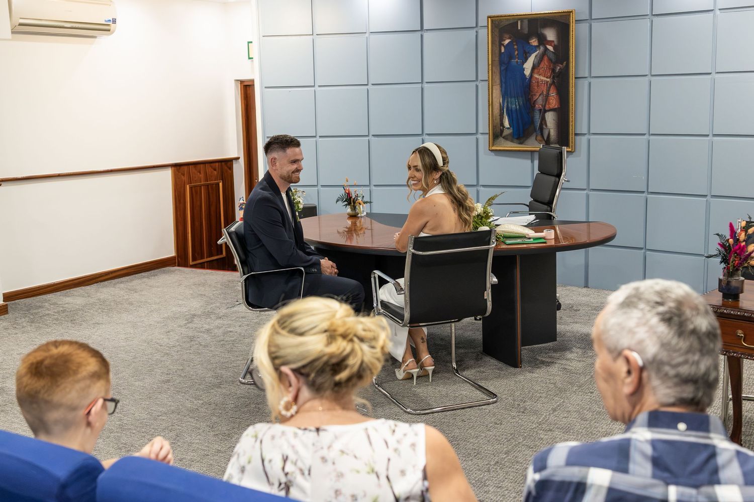Couple signing wedding documents at a registry office ceremony with guests seated in the background.