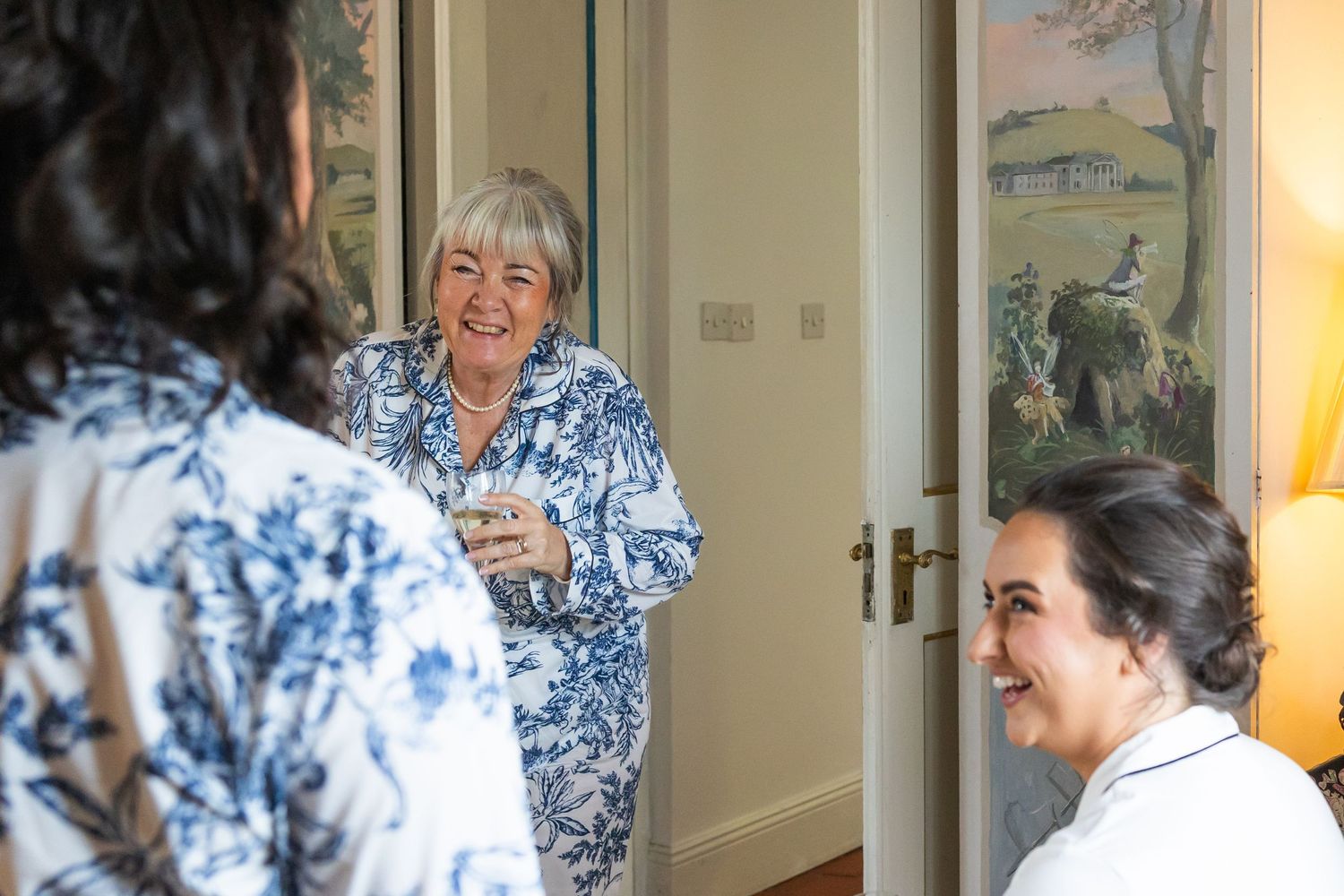 Three women in floral attire share a joyful conversation in an elegantly decorated room with artwork on the walls.