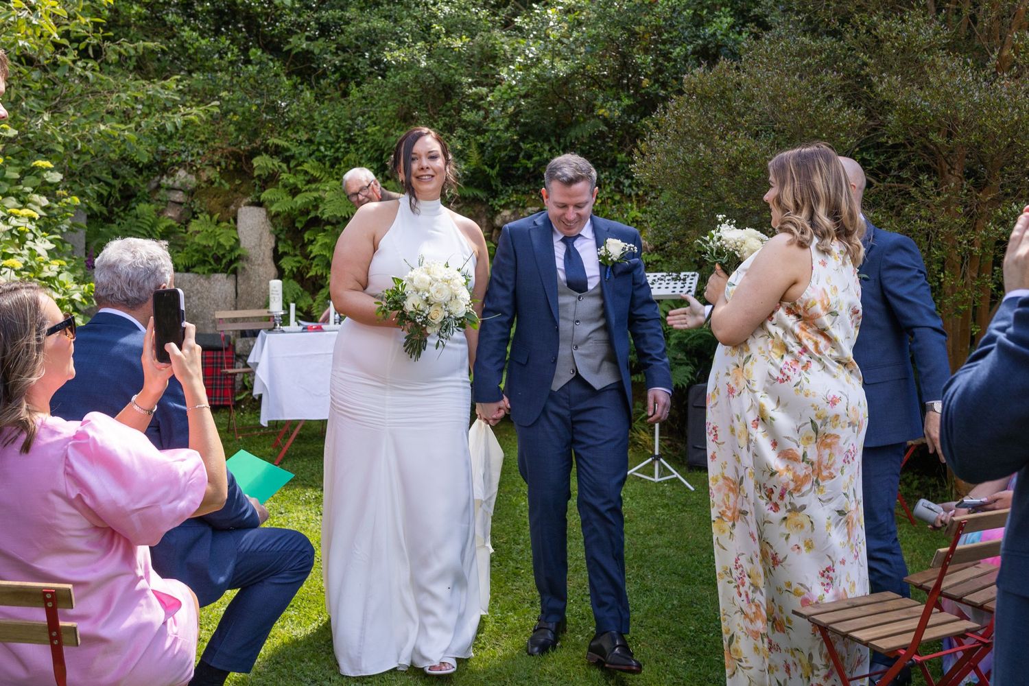 Bride and groom walk hand-in-hand during outdoor garden wedding ceremony surrounded by smiling guests on a sunny day.