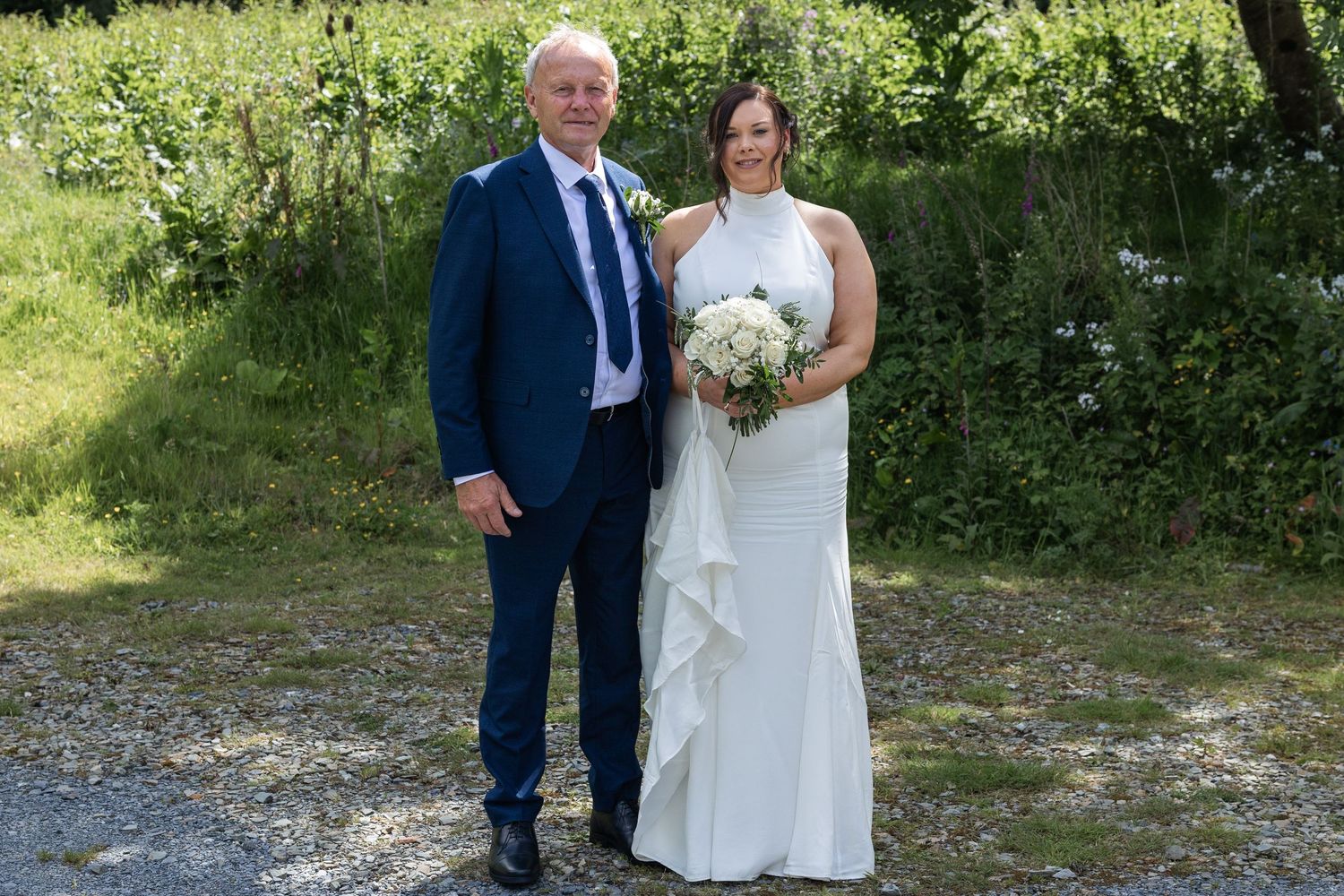 Bride and groom posing outdoors on their wedding day, bride holding white bouquet, groom in navy suit.