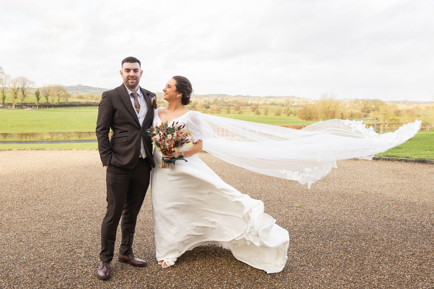 Bride in flowing white gown with veil blowing in wind poses with groom outdoors.