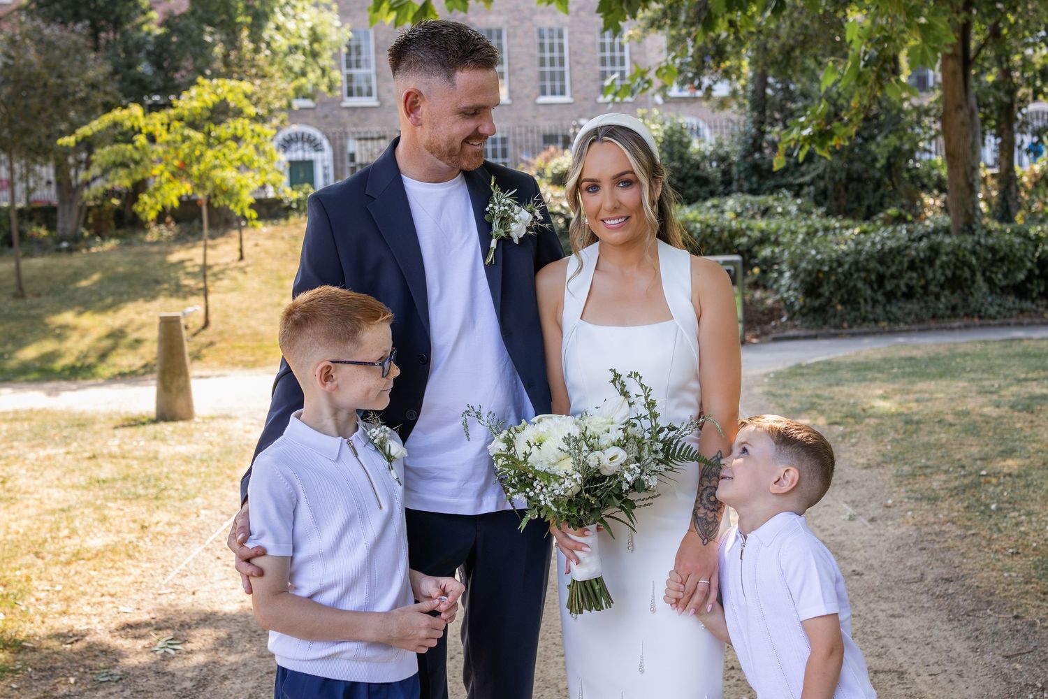 Bride and groom stand with two young boys in a sunny park, bride holding white floral bouquet.