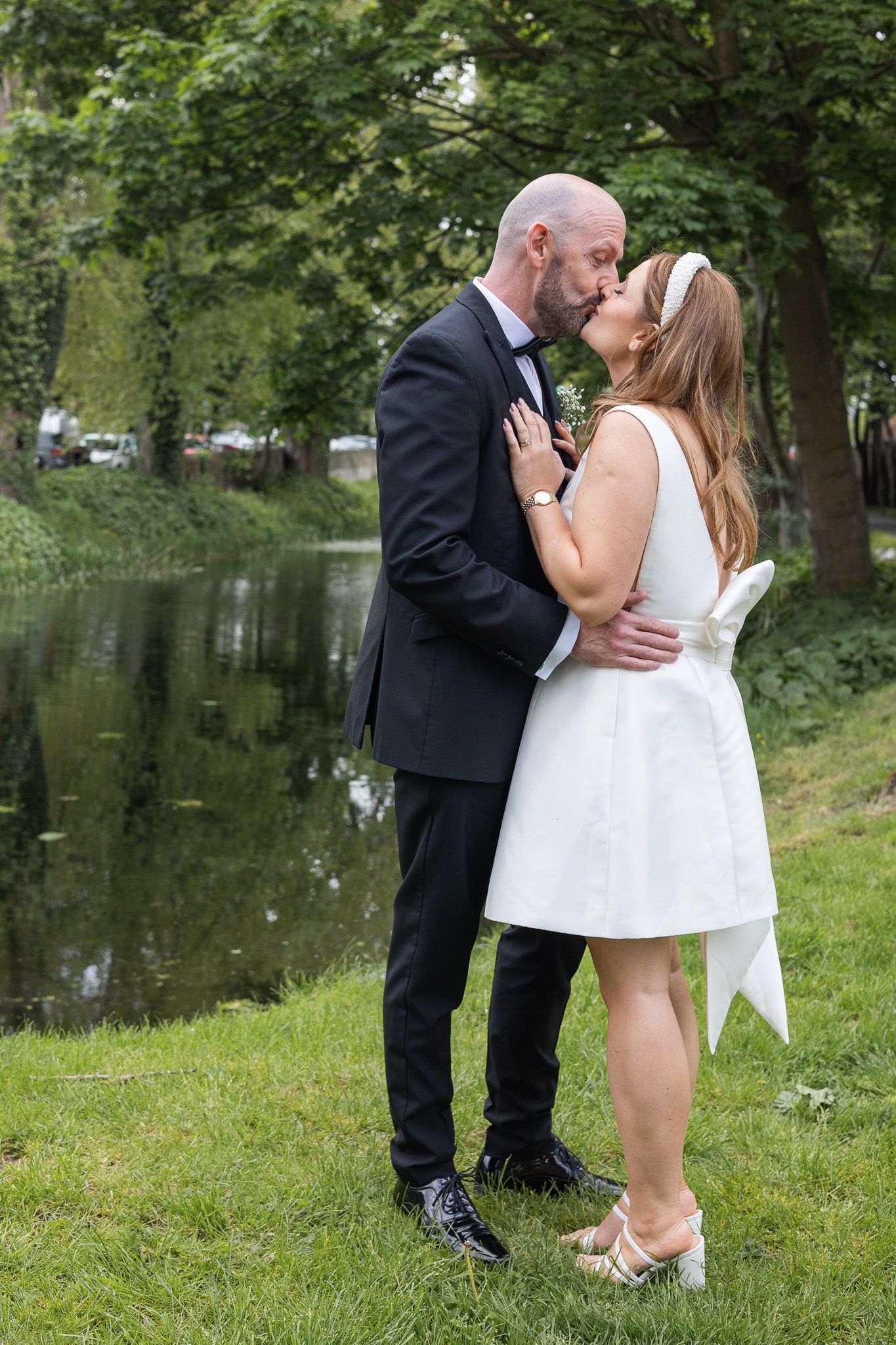 A Dublin couple shares romantic kisses by a scenic pond surrounded by lush green trees.