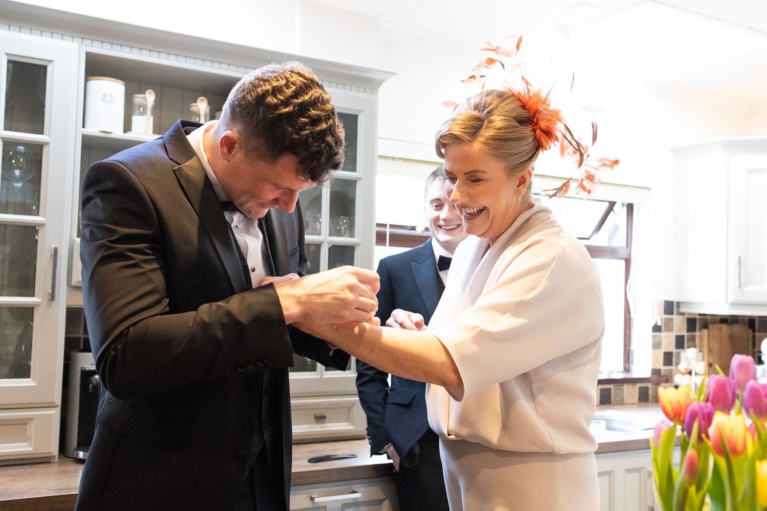 Wedding preparation scene with people helping with formal attire near bright floral arrangements.