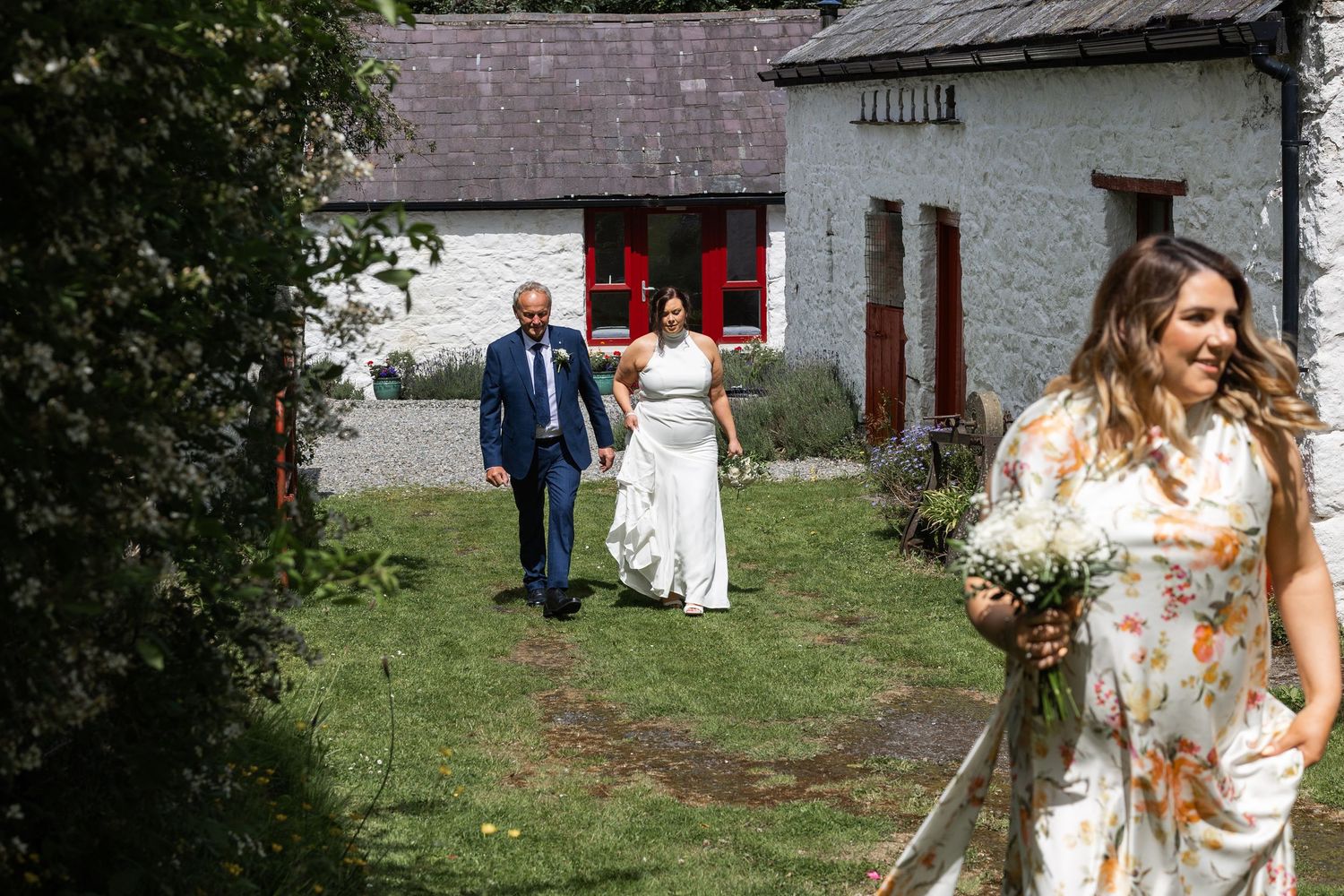 Bride and groom walking hand in hand toward a rustic stone cottage, accompanied by a bridesmaid holding a floral bouquet.