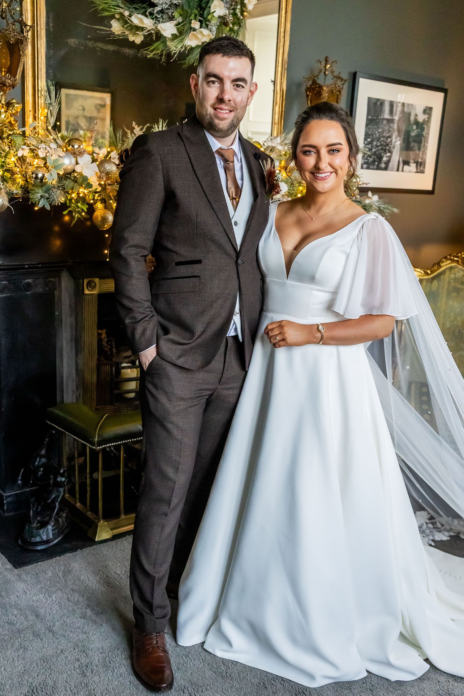 A smiling bride in a white gown and groom in a brown suit pose together indoors near a fireplace.