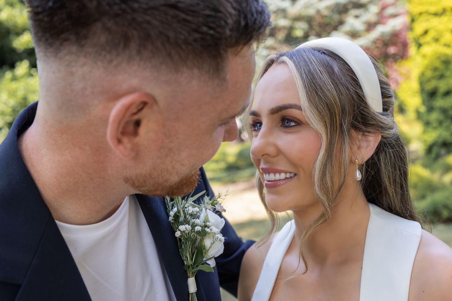 A smiling bride in a white dress and headband gazes at her groom in a navy suit with a floral boutonniere.