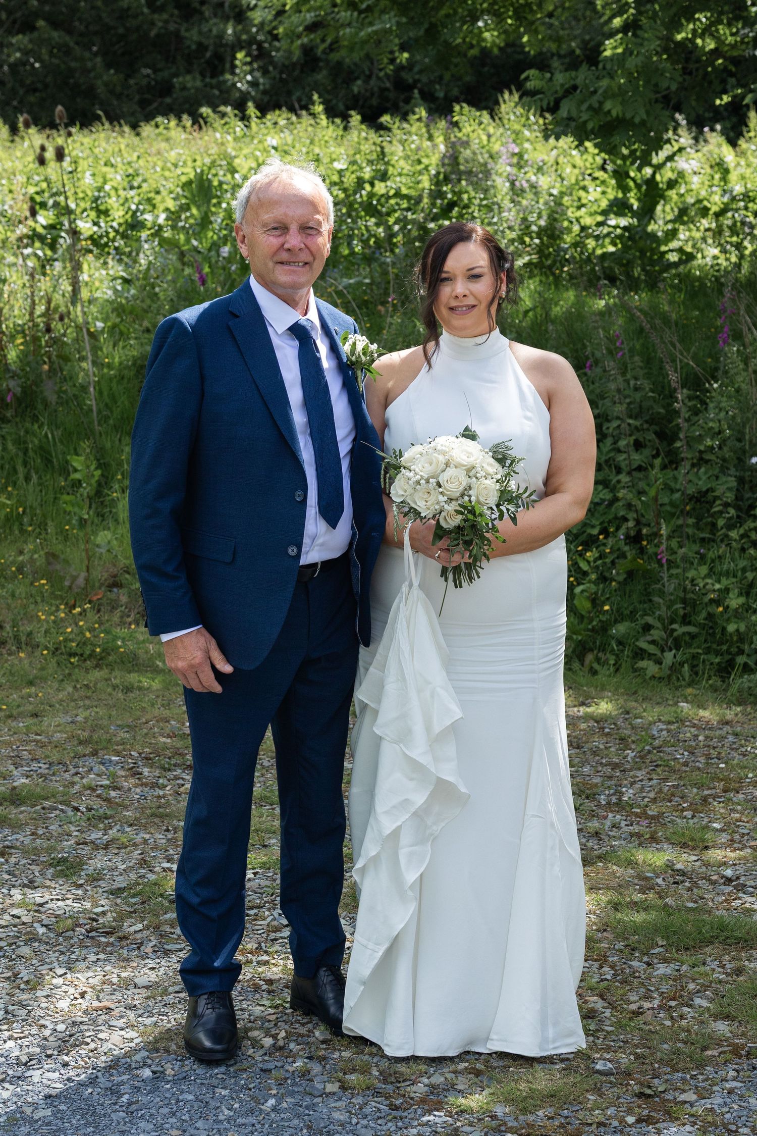 Bride in white halter gown holding bouquet stands with man in navy suit outdoors at wedding.