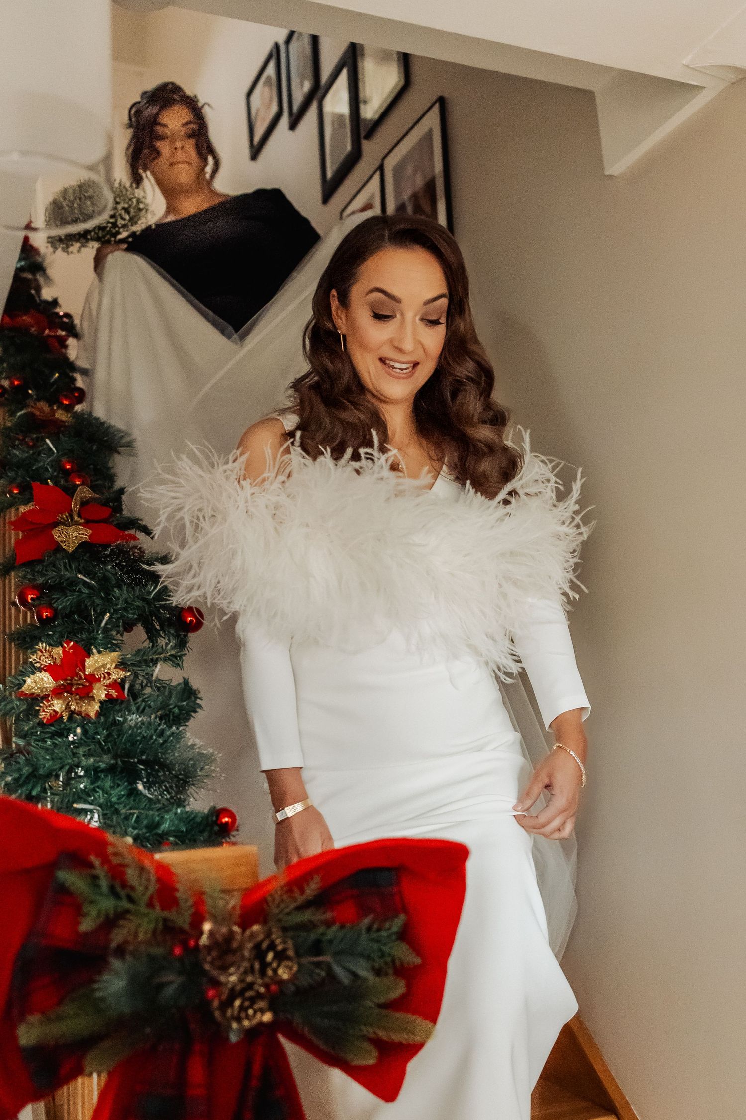 Bride in white feather-trimmed gown smiling beside a decorated Christmas tree with red bows.
