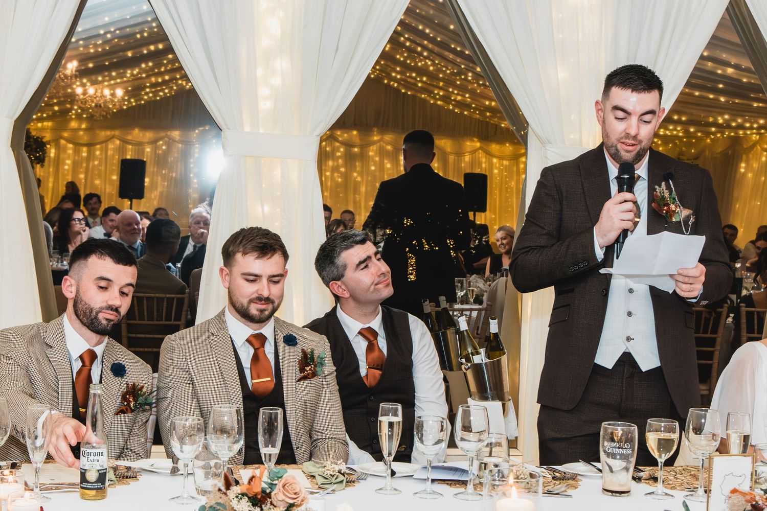 Groomsmen listen as best man delivers wedding speech at elegant marquee reception with fairy lights.