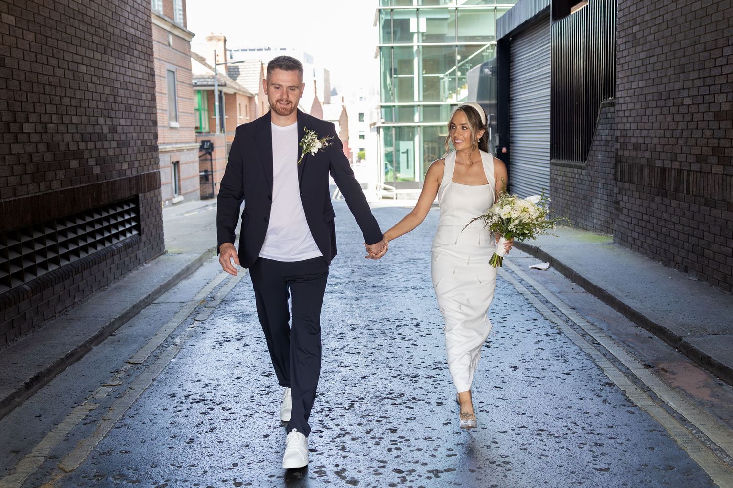 A bride and groom walk hand-in-hand down a cobblestone alley, she in a white dress holding a bouquet, he in a dark suit.