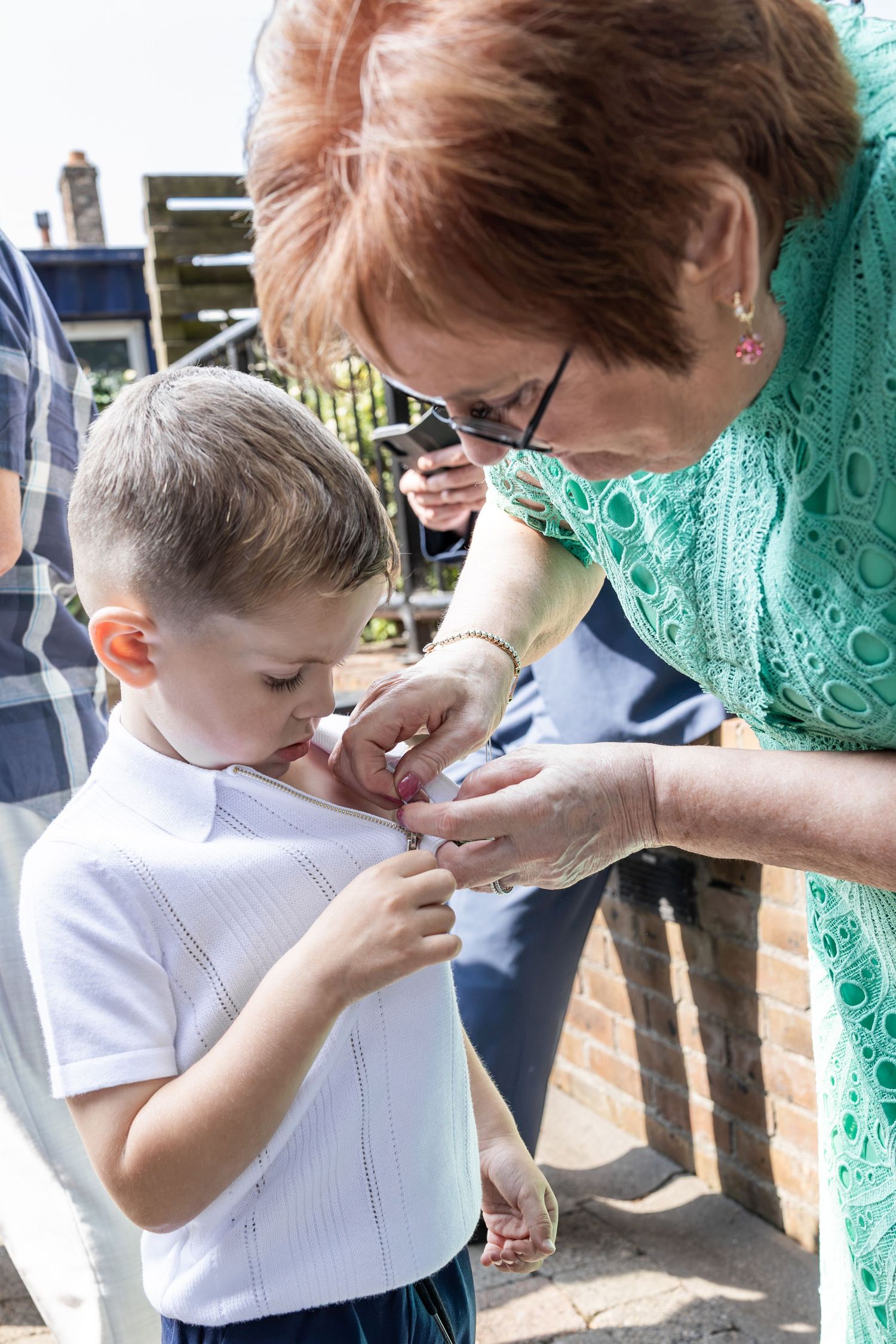 An older woman helps a young boy button his white shirt at an outdoor family gathering.