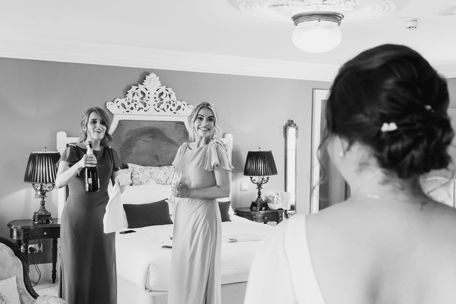 Black and white photo of bride being admired by two bridesmaids in elegant bedroom during wedding preparations.