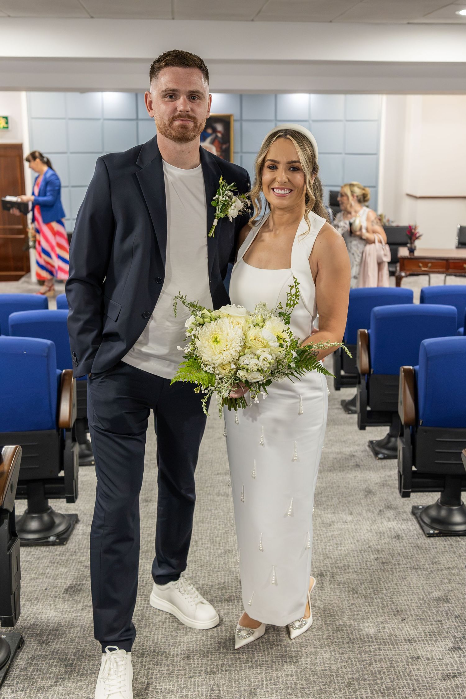 Bride in white dress holding bouquet with groom in navy suit posing in registry office aisle.