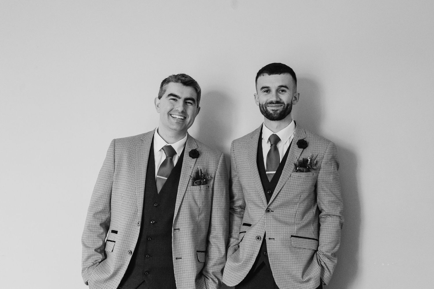 Two grooms in matching striped suits with boutonnieres smiling together in a black and white wedding photo.