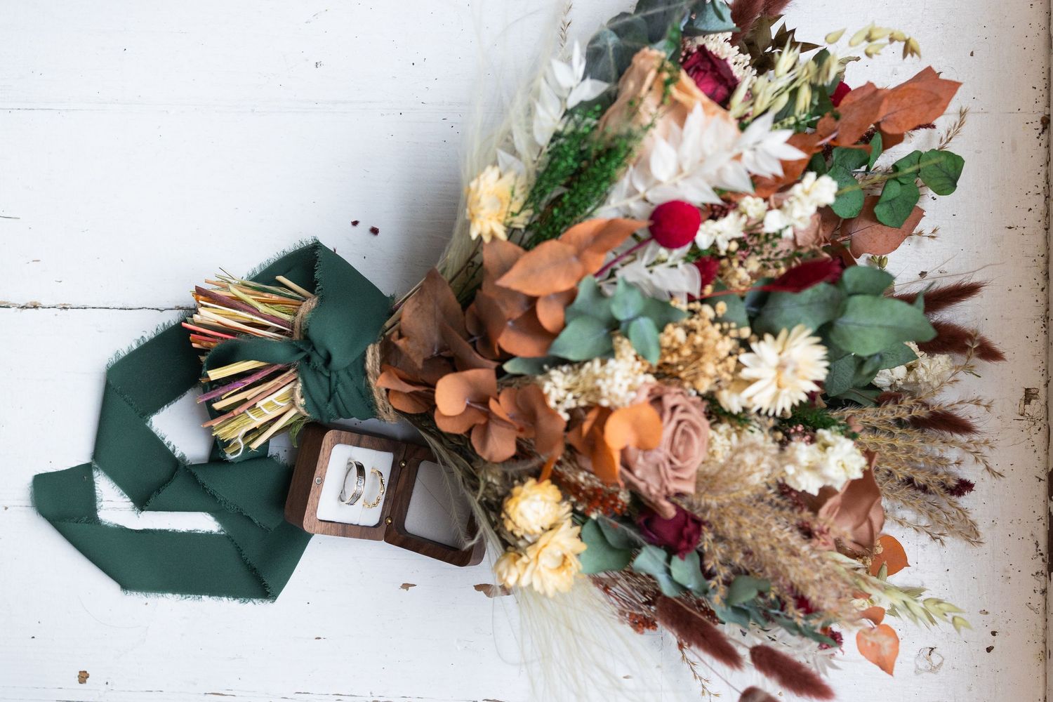Rustic wedding bouquet with dried flowers, eucalyptus, and roses beside a ring box and green ribbon.