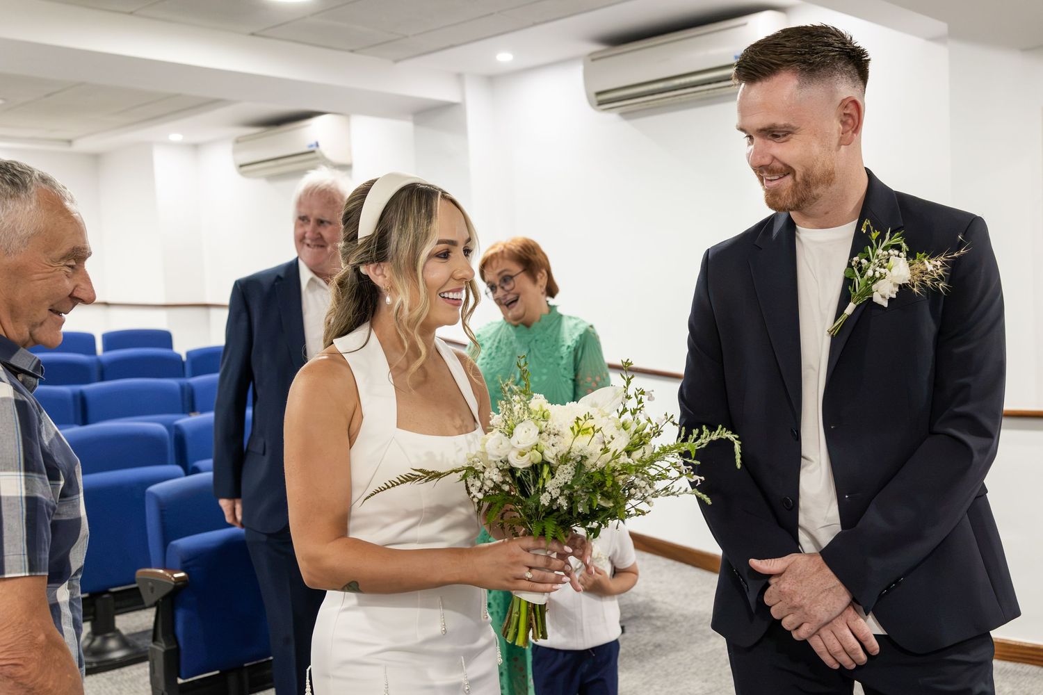 Bride holding white bouquet smiles at groom in navy suit with boutonniere during intimate wedding ceremony.