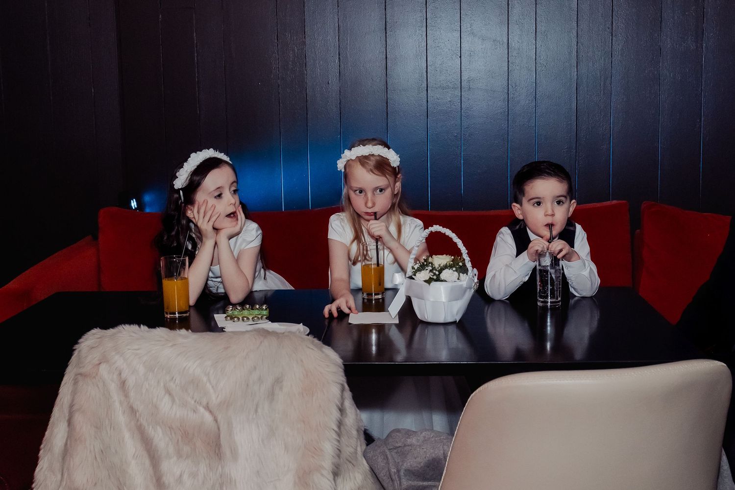 Three children in formal attire sit at a table during a wedding reception, looking bored and tired.