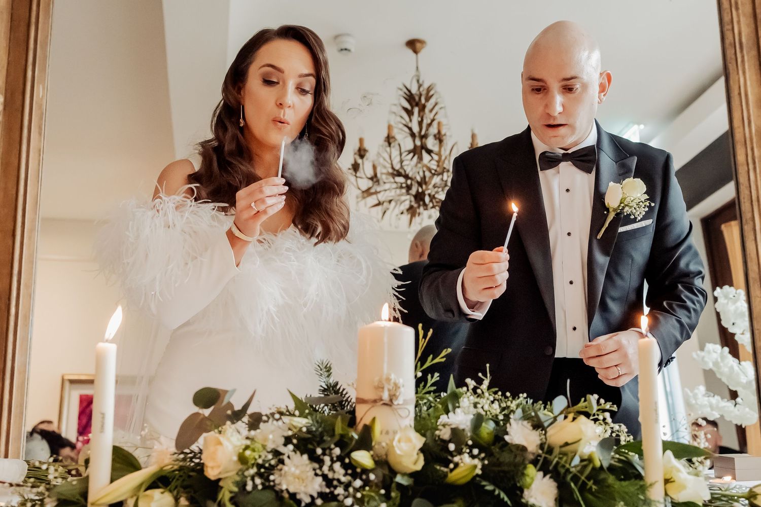 Bride and groom lighting candles at wedding ceremony altar decorated with white flowers and greenery.