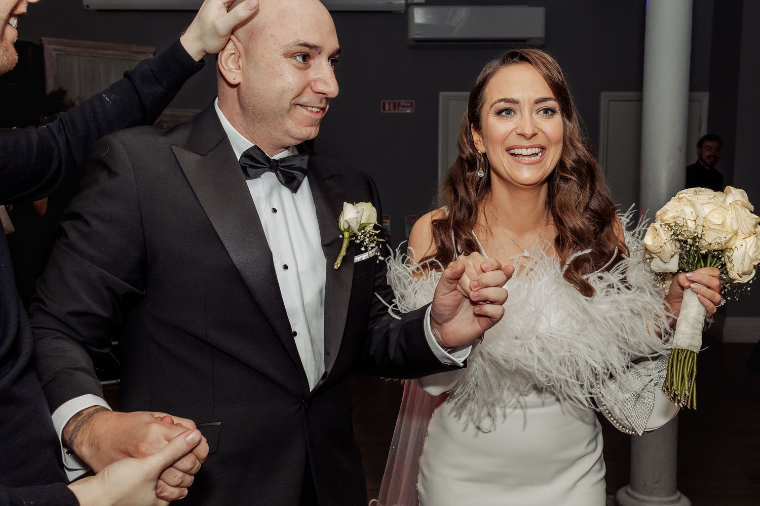 Happy couple shares first dance at wedding reception, bride in white gown with feathers holding bouquet, groom in tuxedo.