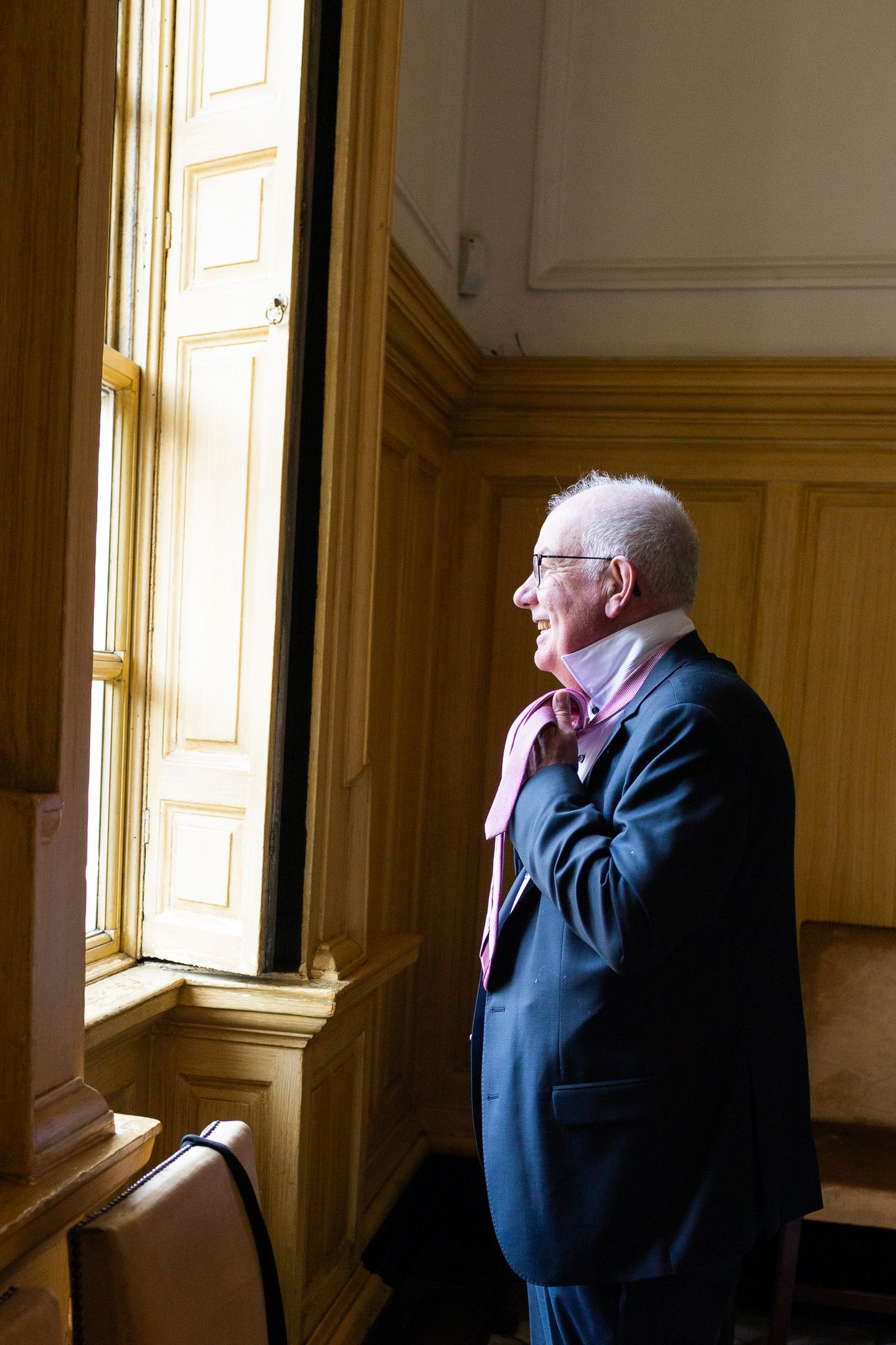 An elderly man in a suit adjusts his tie while standing by a sunlit window in preparations for Dublin wedding.