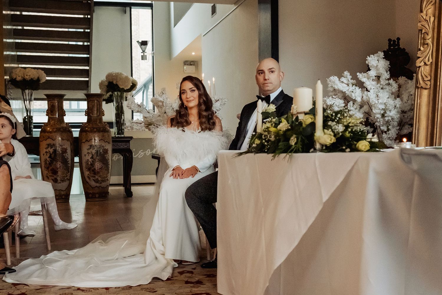 Bride and groom seated at wedding ceremony table adorned with white floral arrangements and candles in elegant venue.