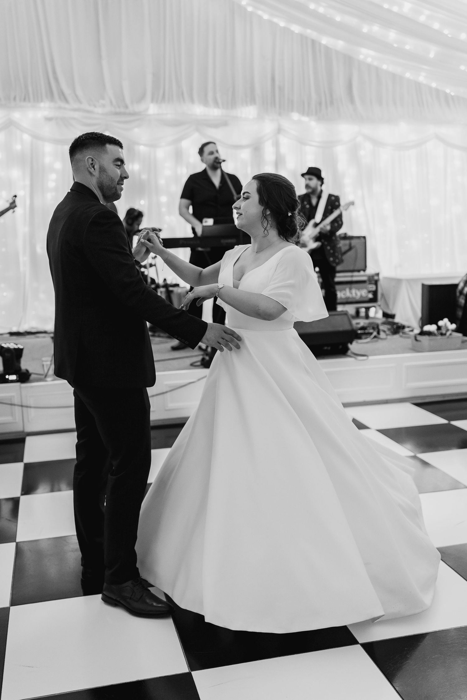 Bride and groom share first dance on checkered floor with live band performing in the background.