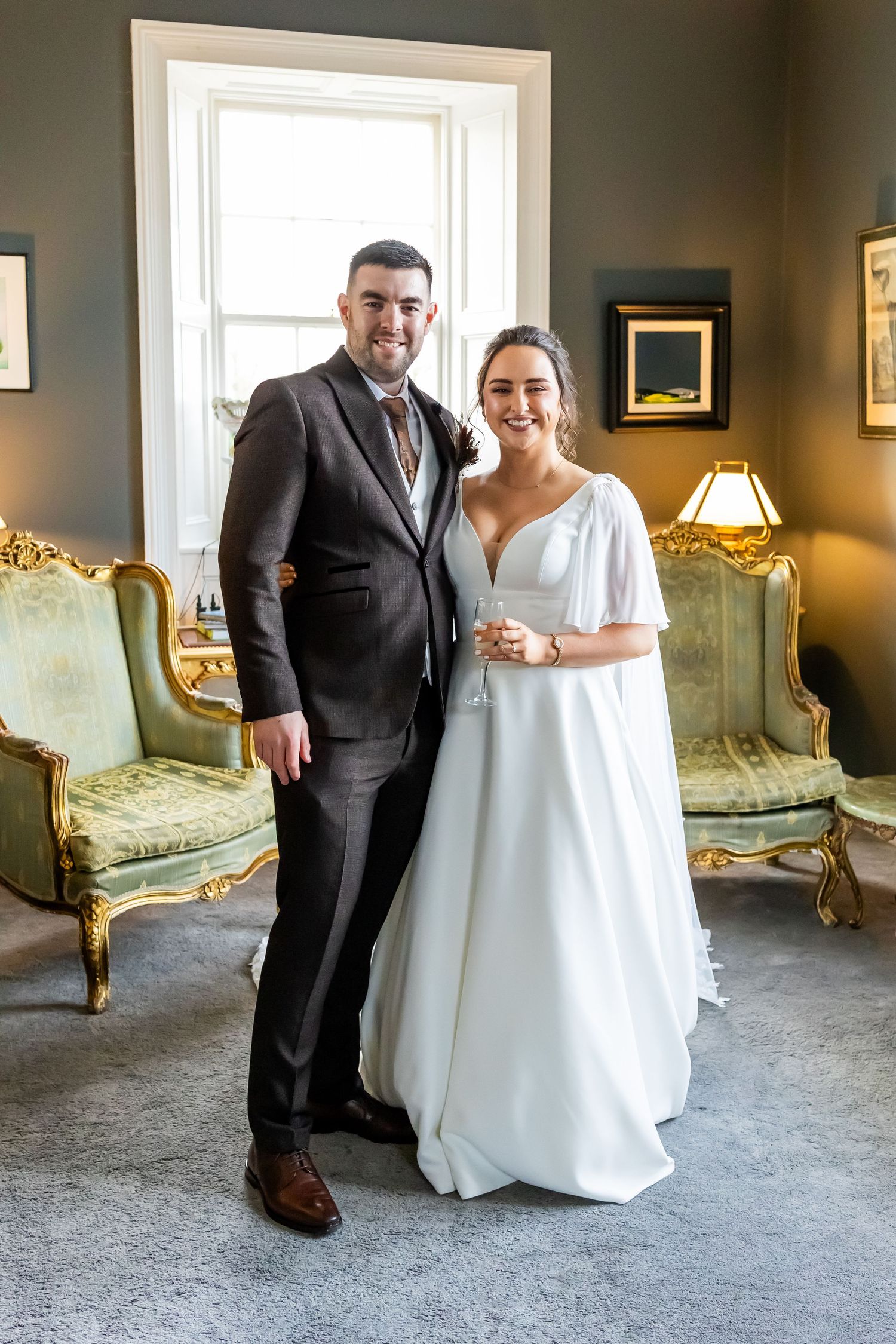 A smiling bride in a white gown and groom in a dark suit pose together in an elegant room with gold furniture.