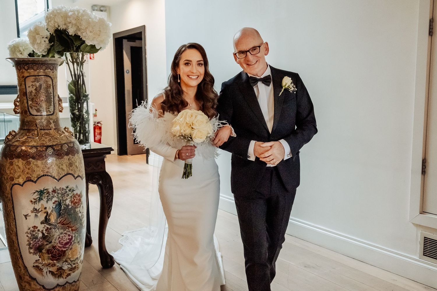 Bride in elegant white gown walks with older gentleman in tuxedo holding bouquet at wedding.