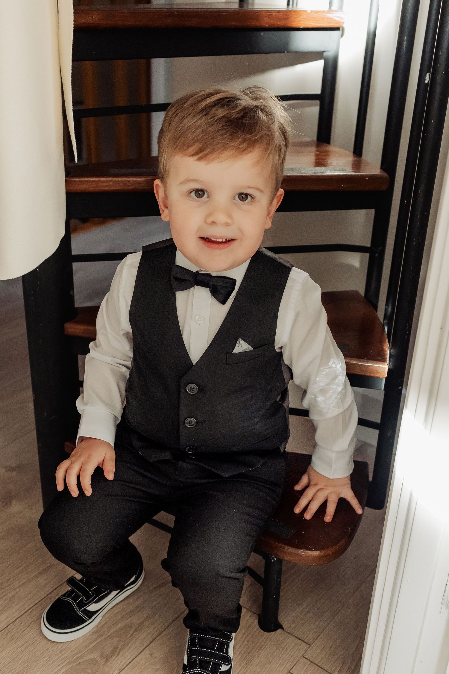 Small child in formal black vest and bowtie sitting on wooden furniture.