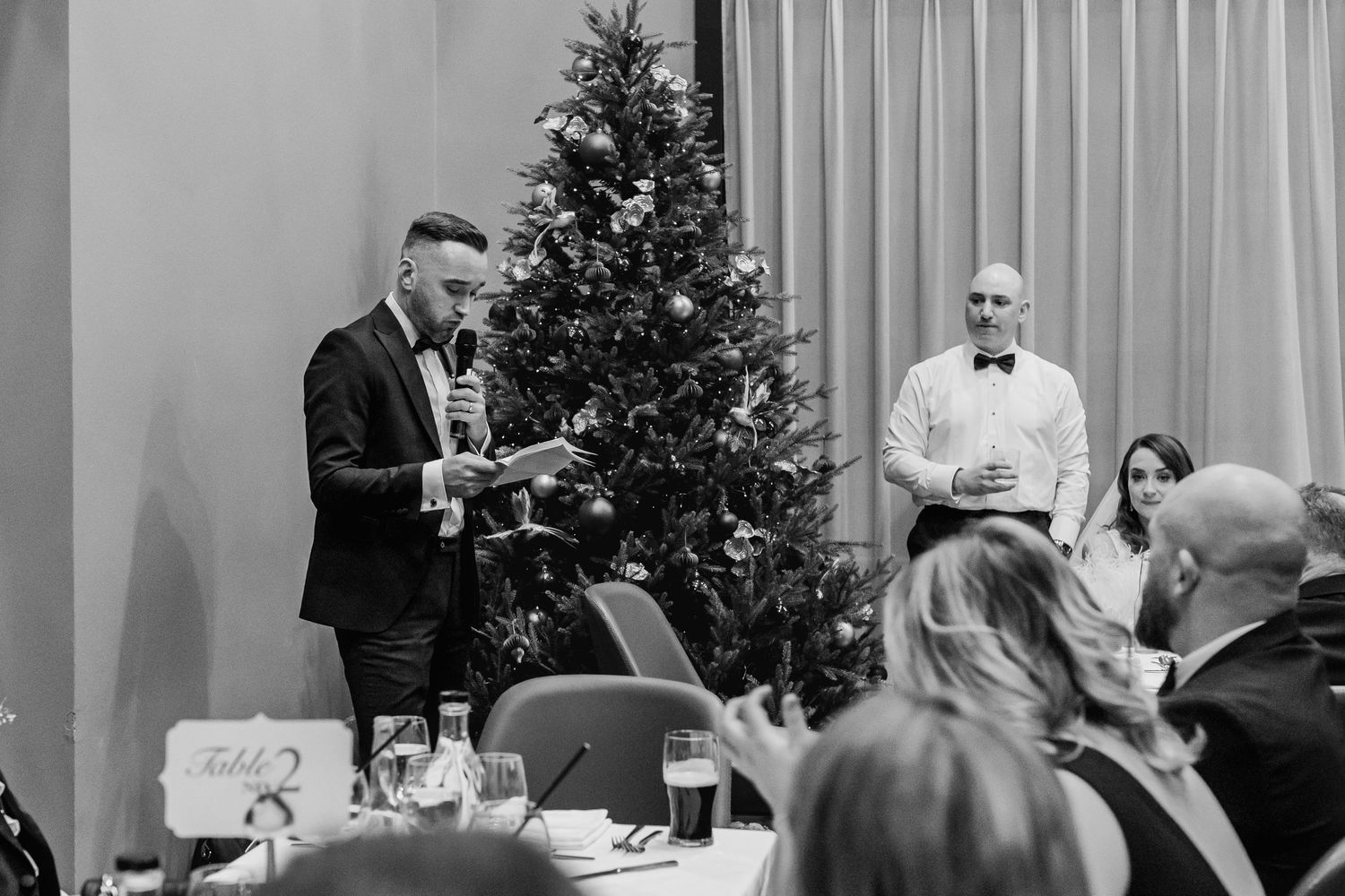 Black and white photo of wedding reception speeches with holiday decor visible.