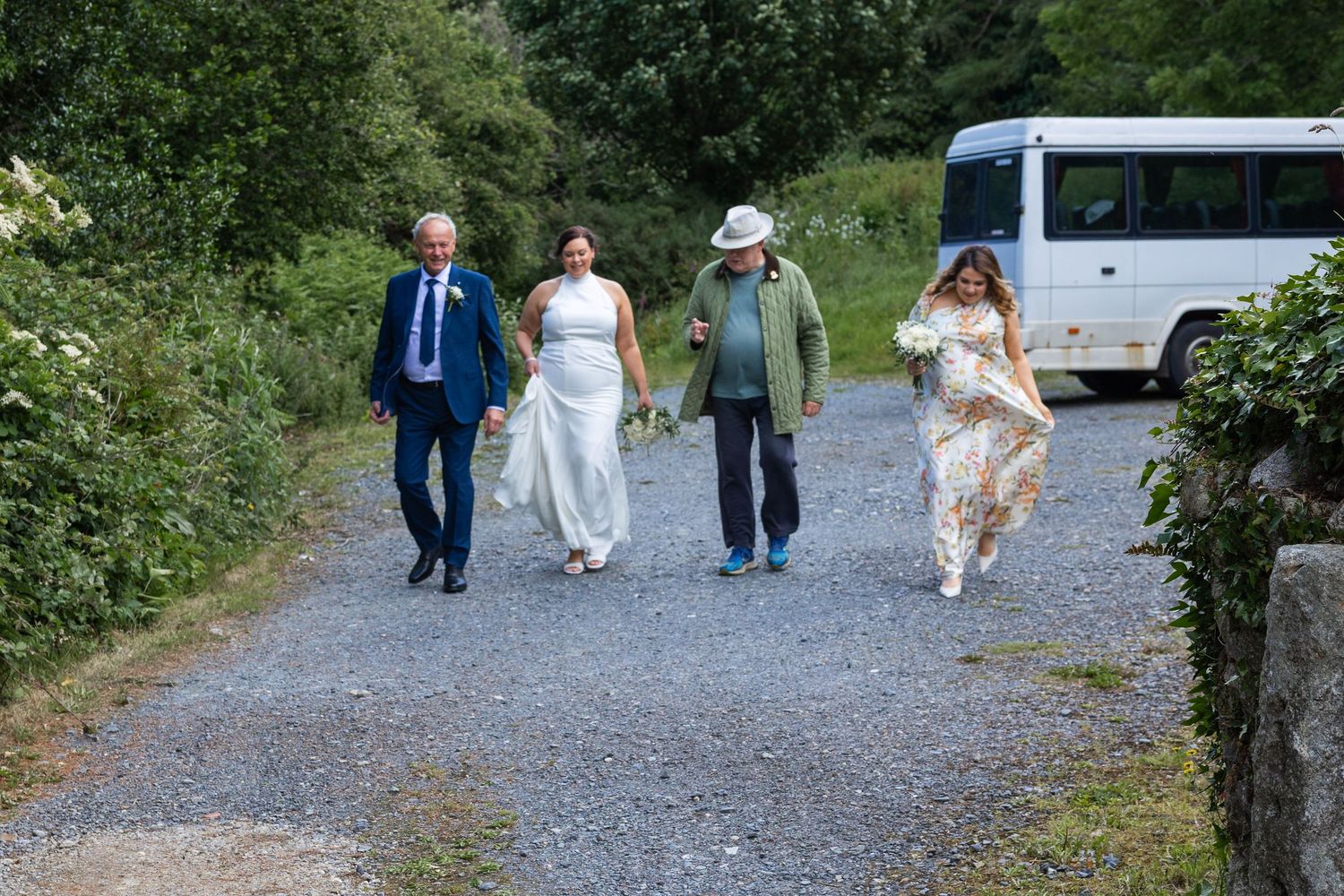 Wedding party walking down a gravel path outdoors, bride in white gown with guests in formal attire near a white bus.