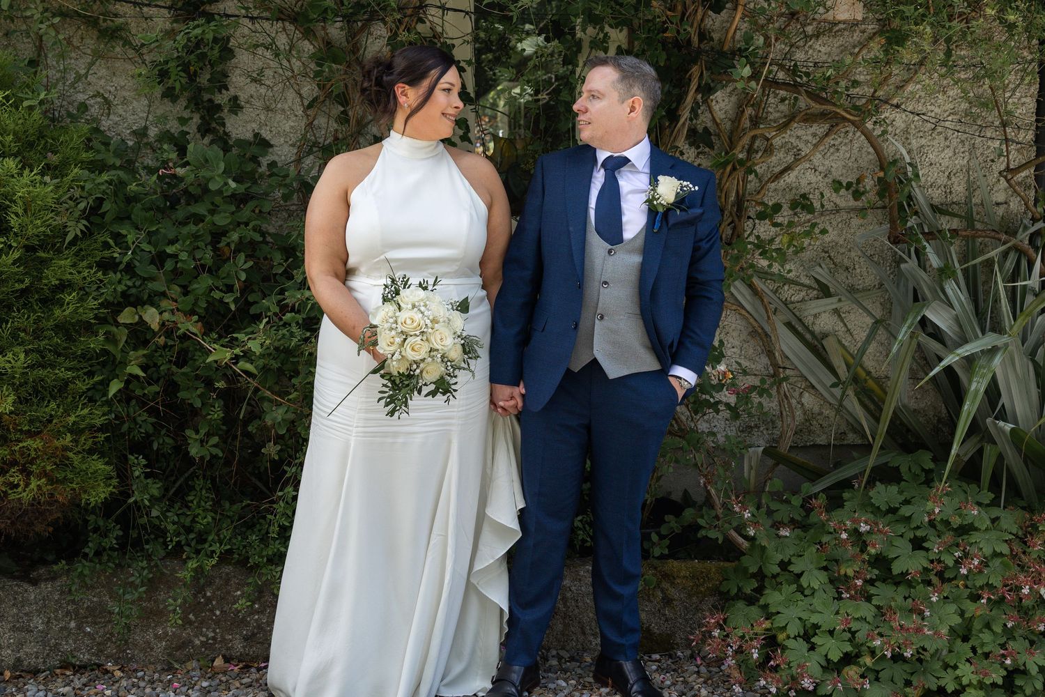 Bride in white halter gown holding bouquet stands with groom in navy suit outdoors at elegant wedding venue.