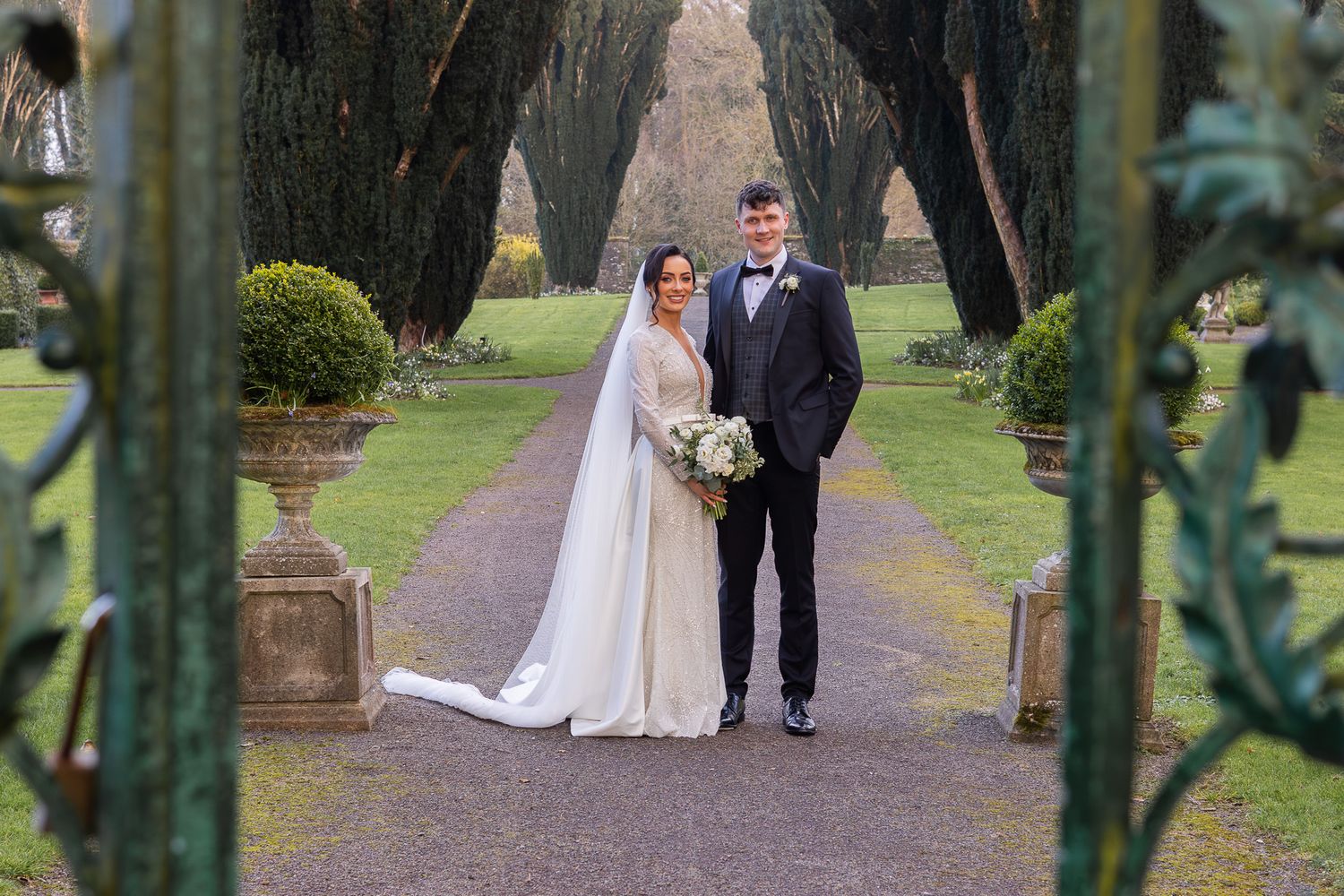 A tree-lined pathway with ornate iron gates creates a natural archway for wedding photos.
