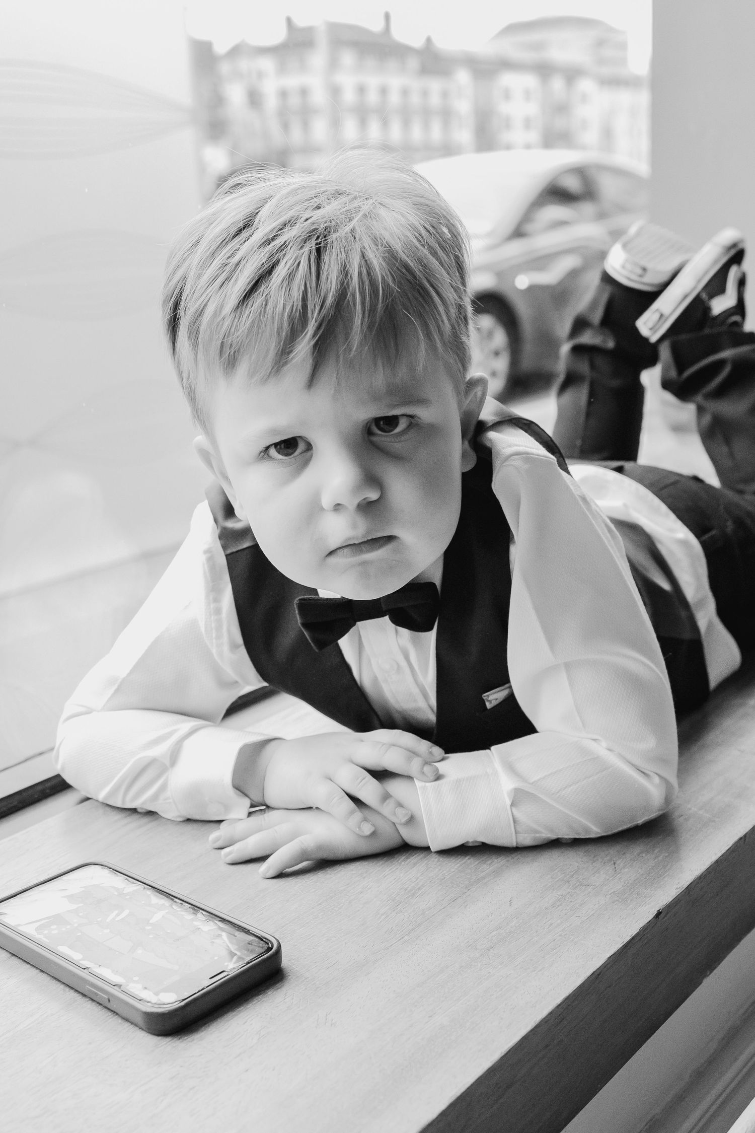 Black and white photo of a young boy in a bow tie and vest lying on a windowsill next to a smartphone.
