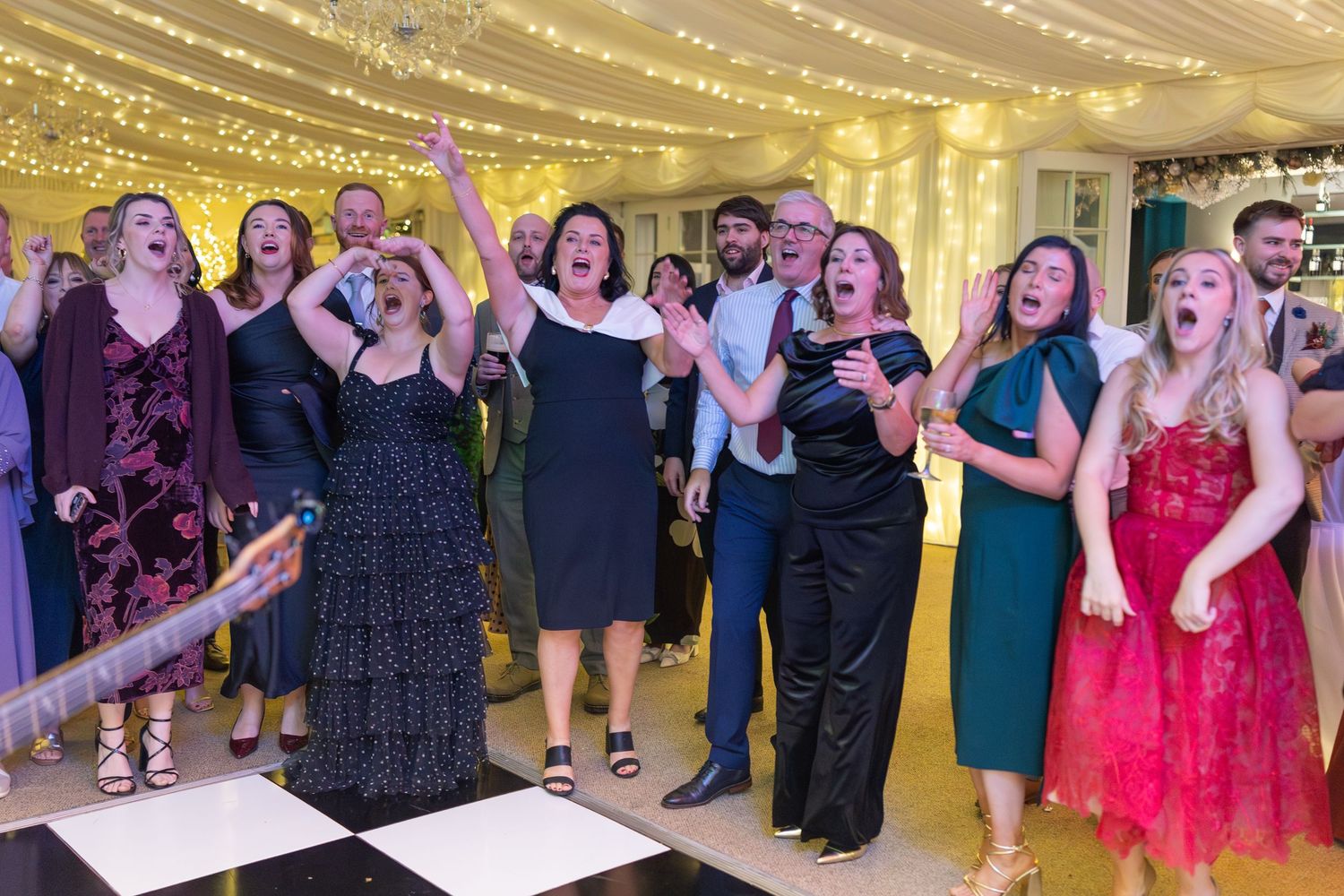 Guests singing and dancing joyfully on a checkered dance floor at a wedding reception under fairy lights.