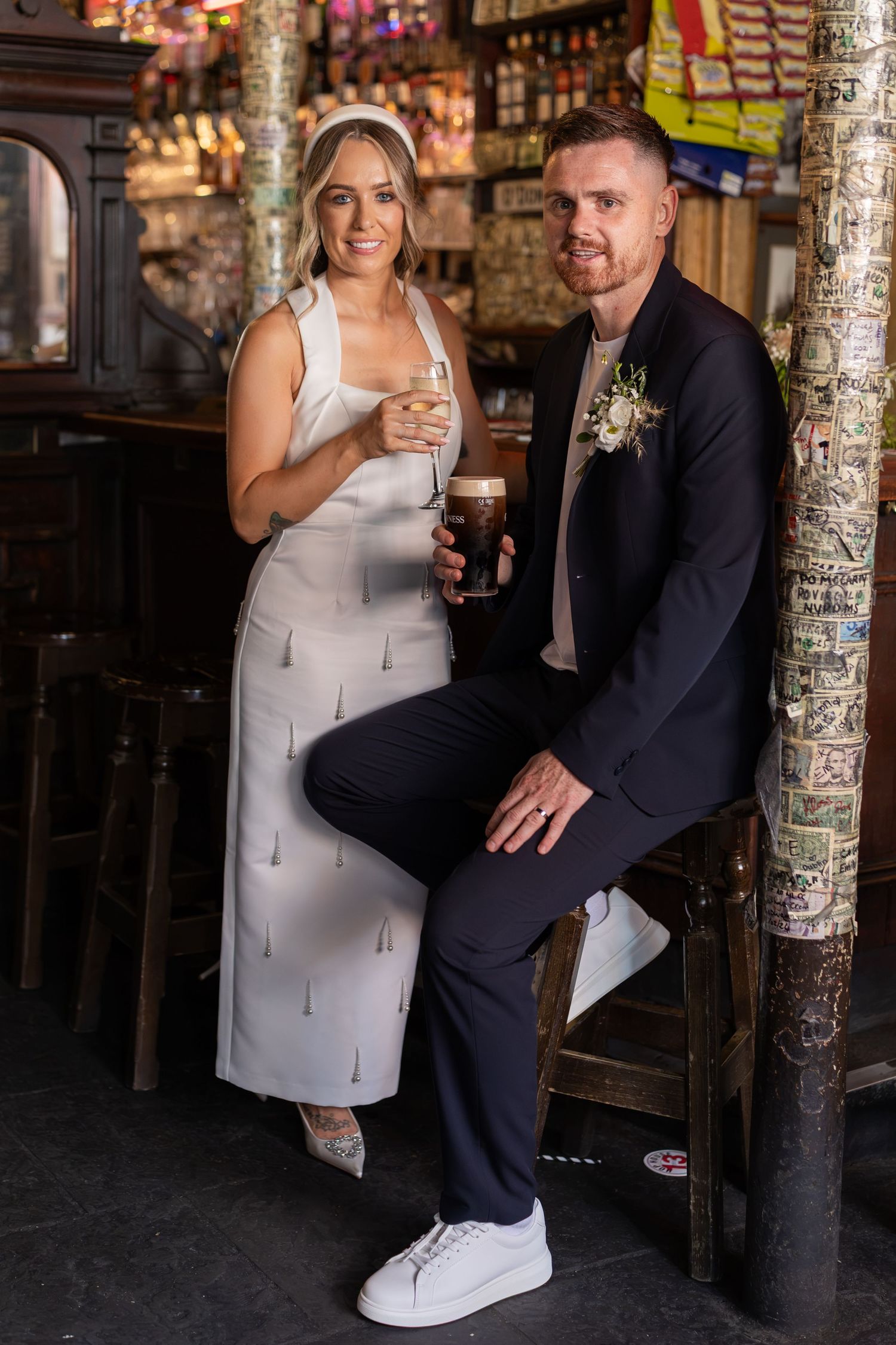 Bride and groom posing in a pub bar, holding drinks, wearing wedding attire with floral boutonniere.